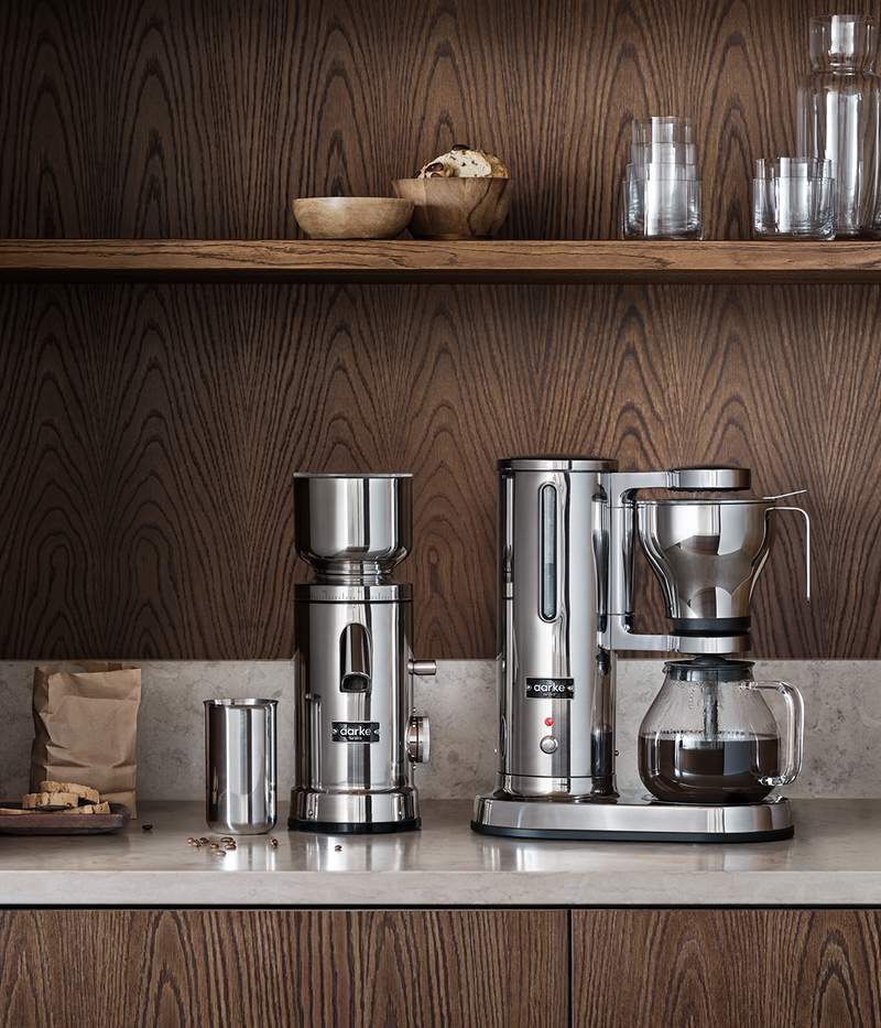 aarke coffee maker and coffee grinder standing on kitchen countertop in a setting of dark wood. 